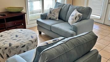 A living room with a grey sofa, patterned ottoman, and a television on the wall.