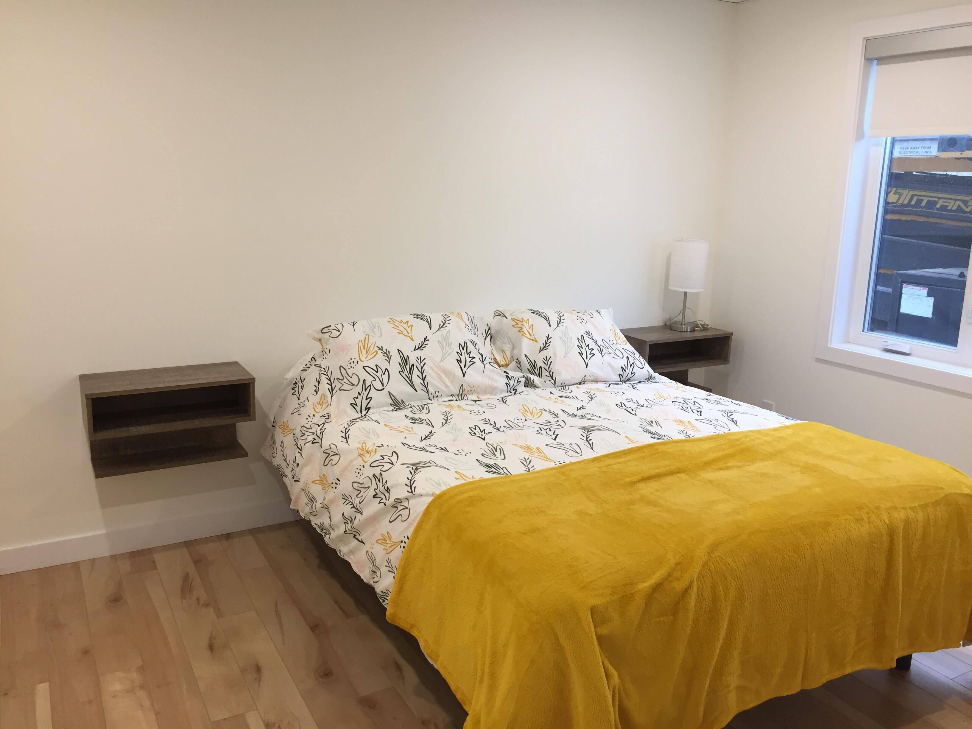A bedroom with a bed, a nightstand, a bedside table, a window, and a shelf.