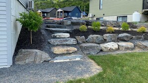 A residential area with a stone pathway leading to houses, surrounded by greenery and a hilly backdrop.