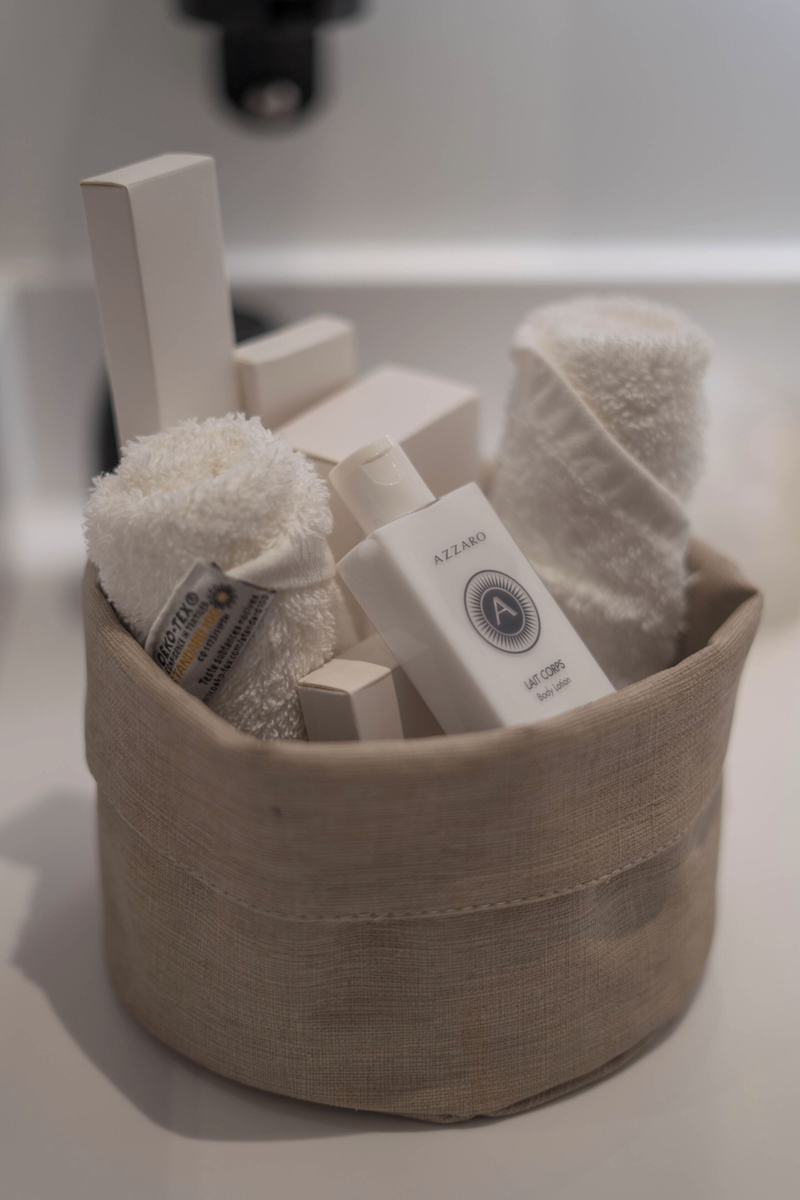 A beige fabric basket containing various toiletries including a white towel, a bottle labeled "AZZARO BODY OIL," and a white bar of soap.