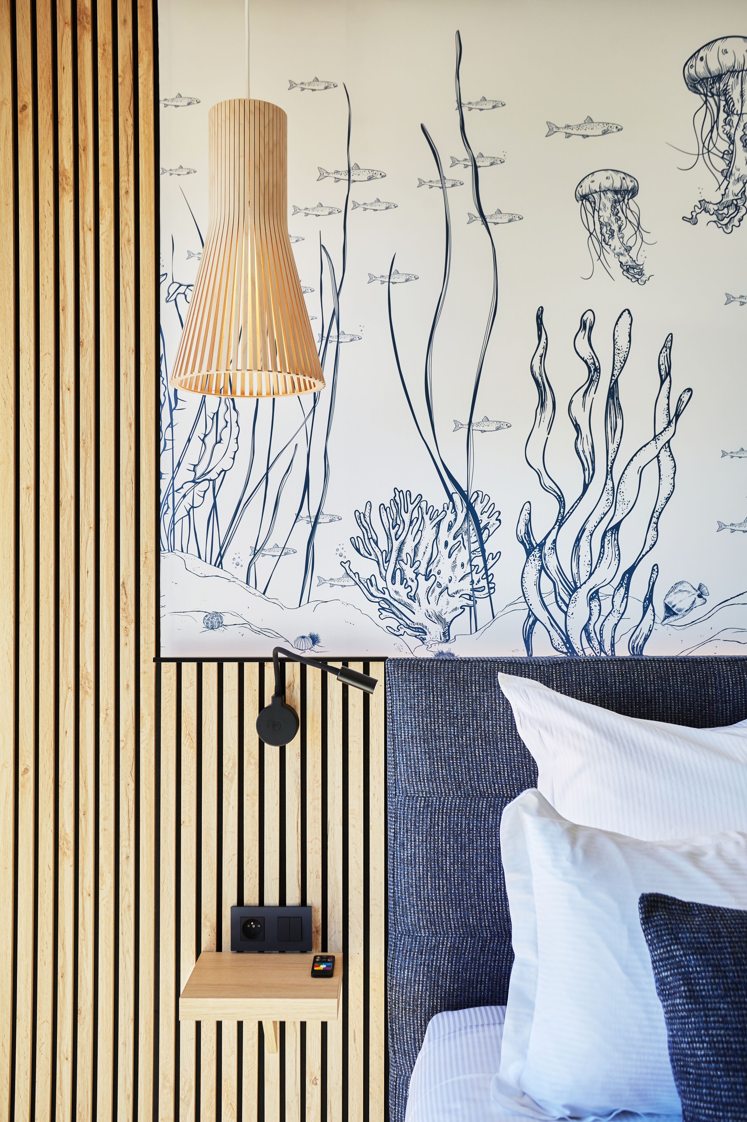 Une pièce avec un mur recouvert de lambris en bois, une suspension lumineuse, une petite étagère avec un haut-parleur et un smartphone, et un lit avec des oreillers blancs.