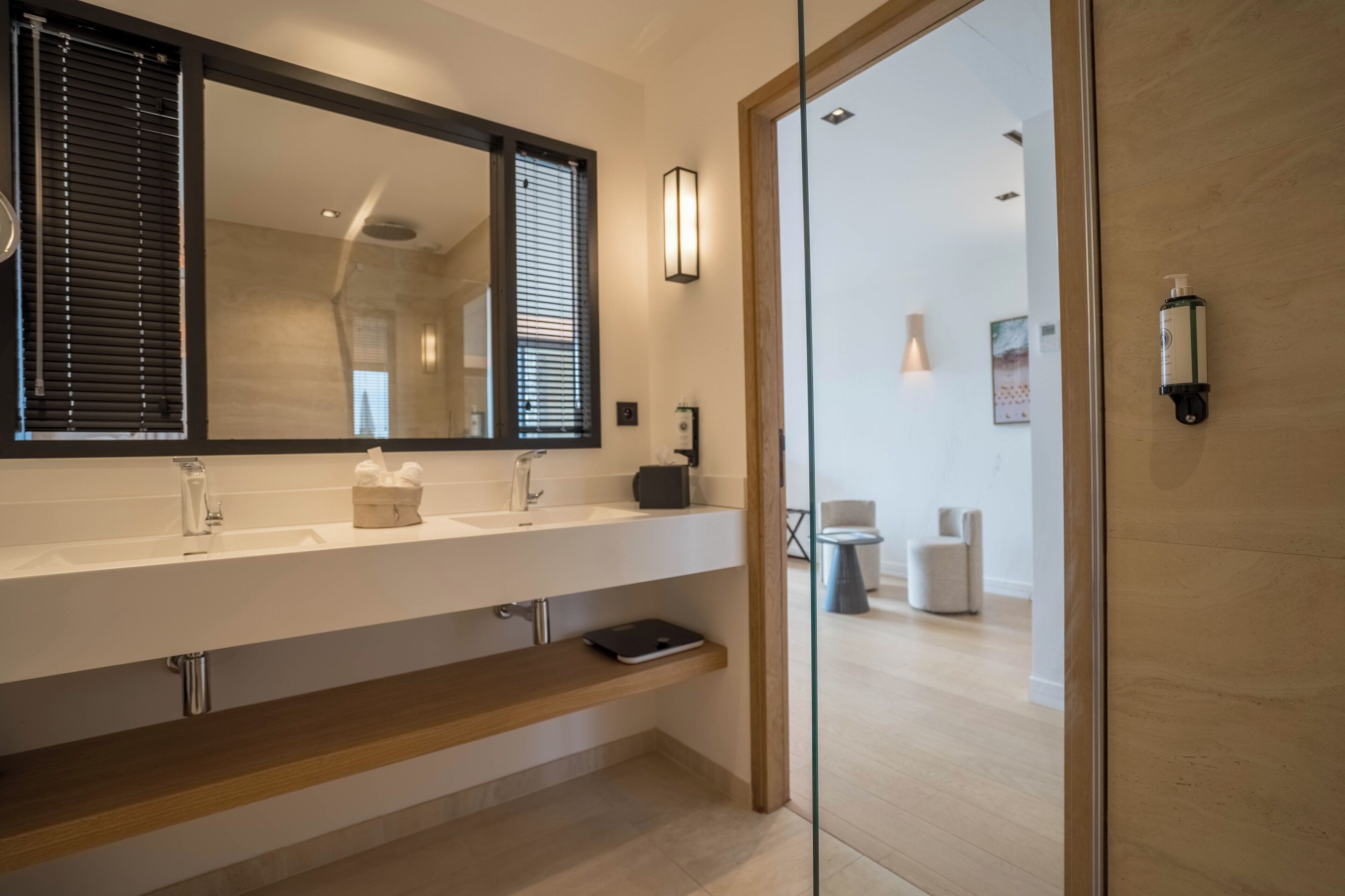 Ein modernes Badezimmer mit einem großen Spiegel, zwei Waschbecken, einer Bank und einer Glastür zum Duschbereich.