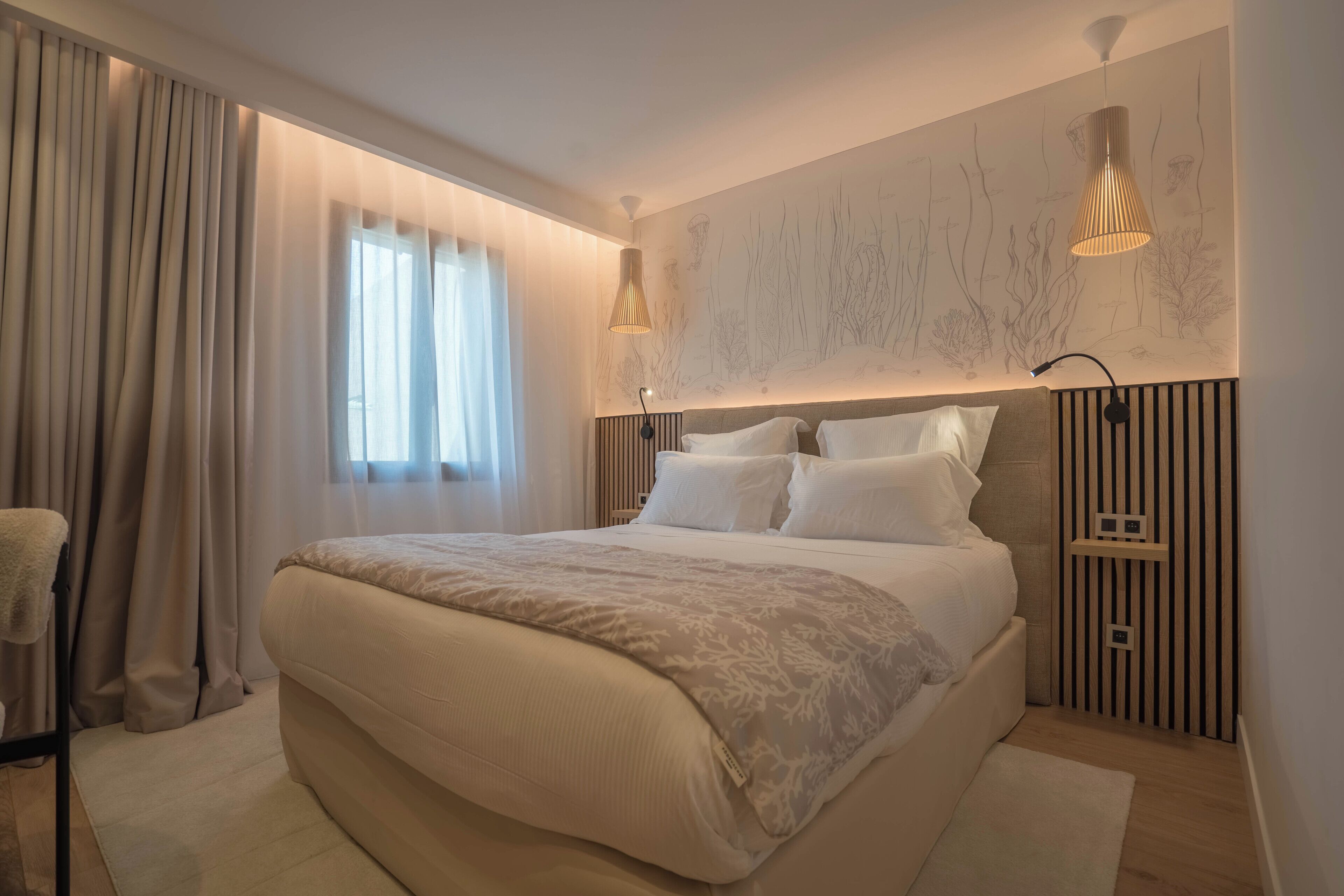A modern bedroom with a large bed, a bedside table, a wall-mounted lamp, and a window with curtains.