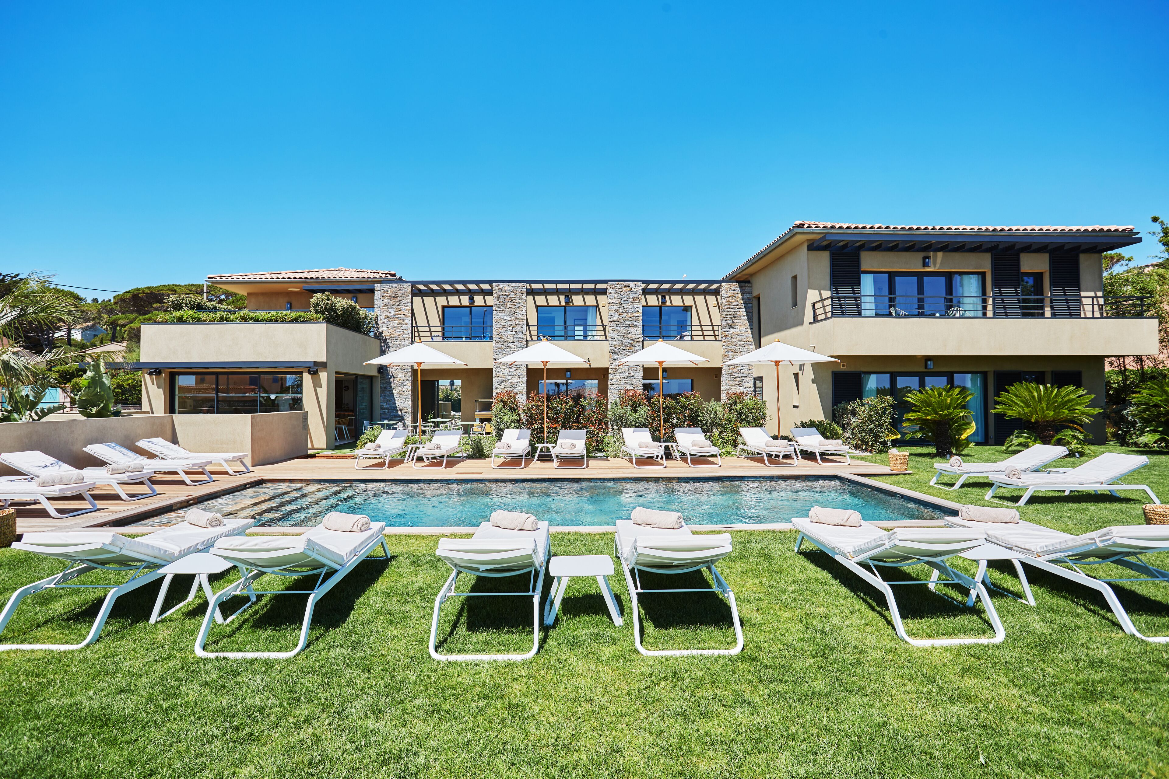 A poolside area with multiple lounge chairs and umbrellas, a clear blue sky, and a modern building with a pool.