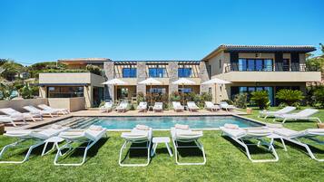 A poolside area with multiple lounge chairs and umbrellas, a clear blue sky, and a modern building with a pool.
