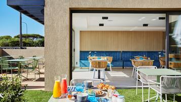 Un espacio de comedor al aire libre con mesas y sillas, una disposición tipo bufé y vistas al interior a través de un vidrio.