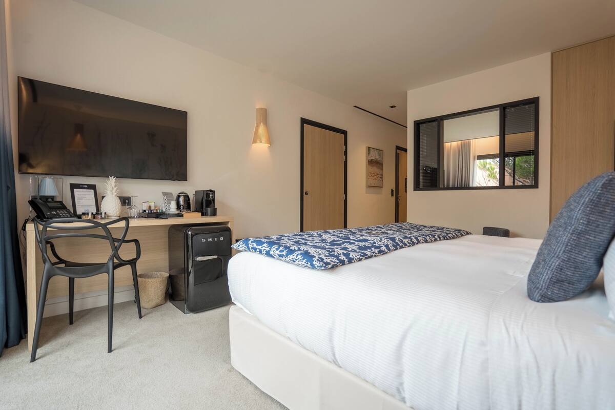 A modern hotel room with a large bed, a desk with a computer, a chair, and a small kitchenette.