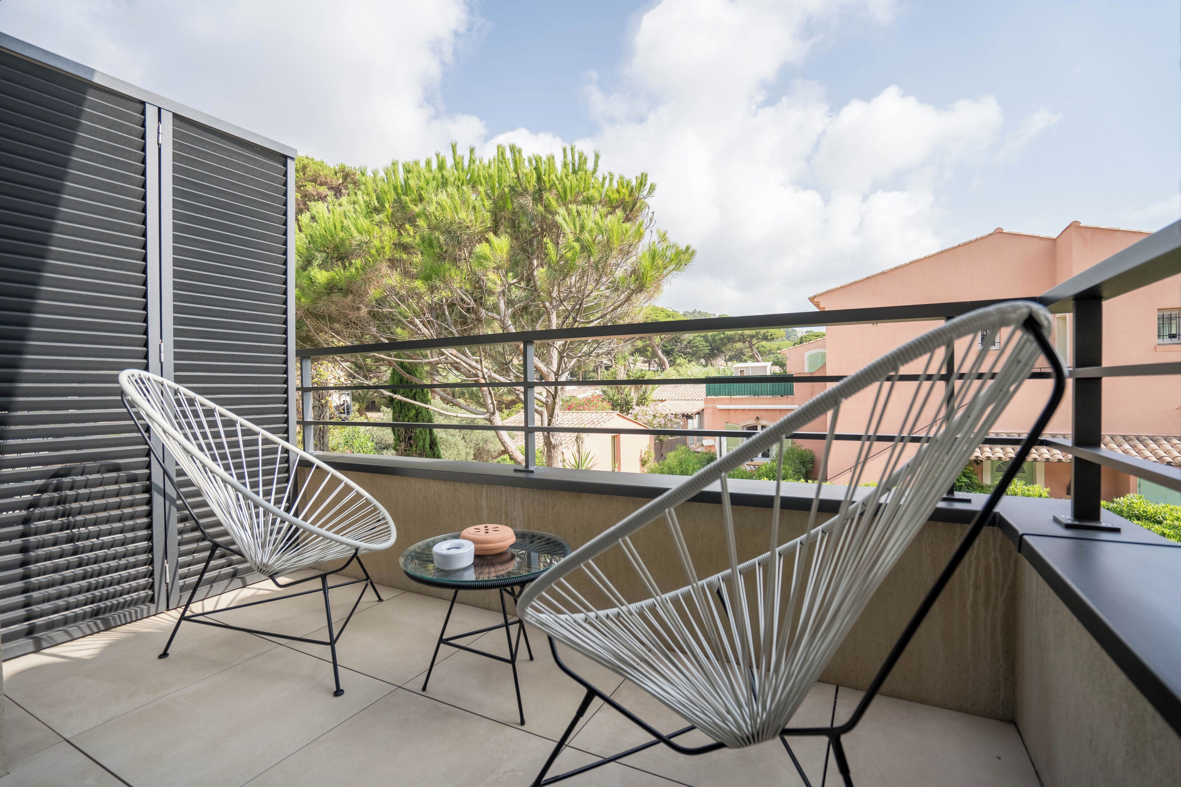 A balcony with two white wire-framed chairs, a small round table with a cup and saucer, and a view of buildings and trees in the background.