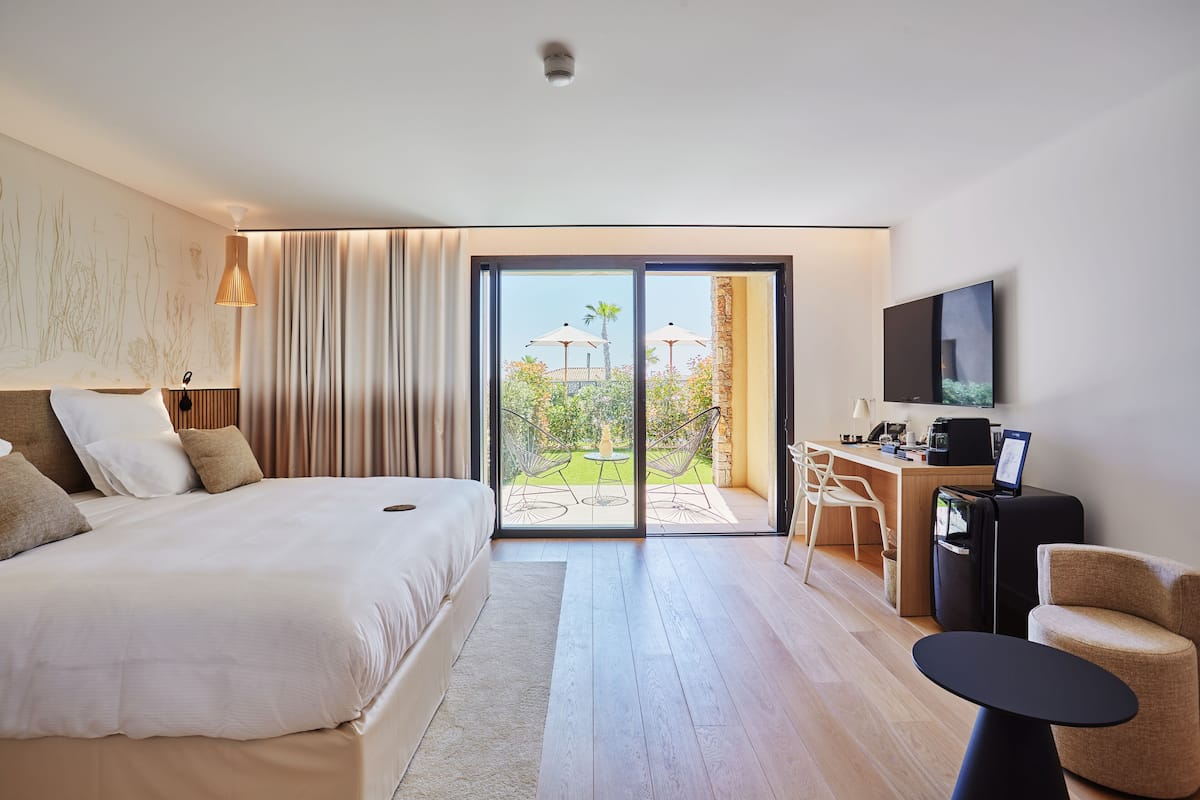 A modern hotel room with a large bed, a desk with a computer, a TV, and a balcony with outdoor furniture.