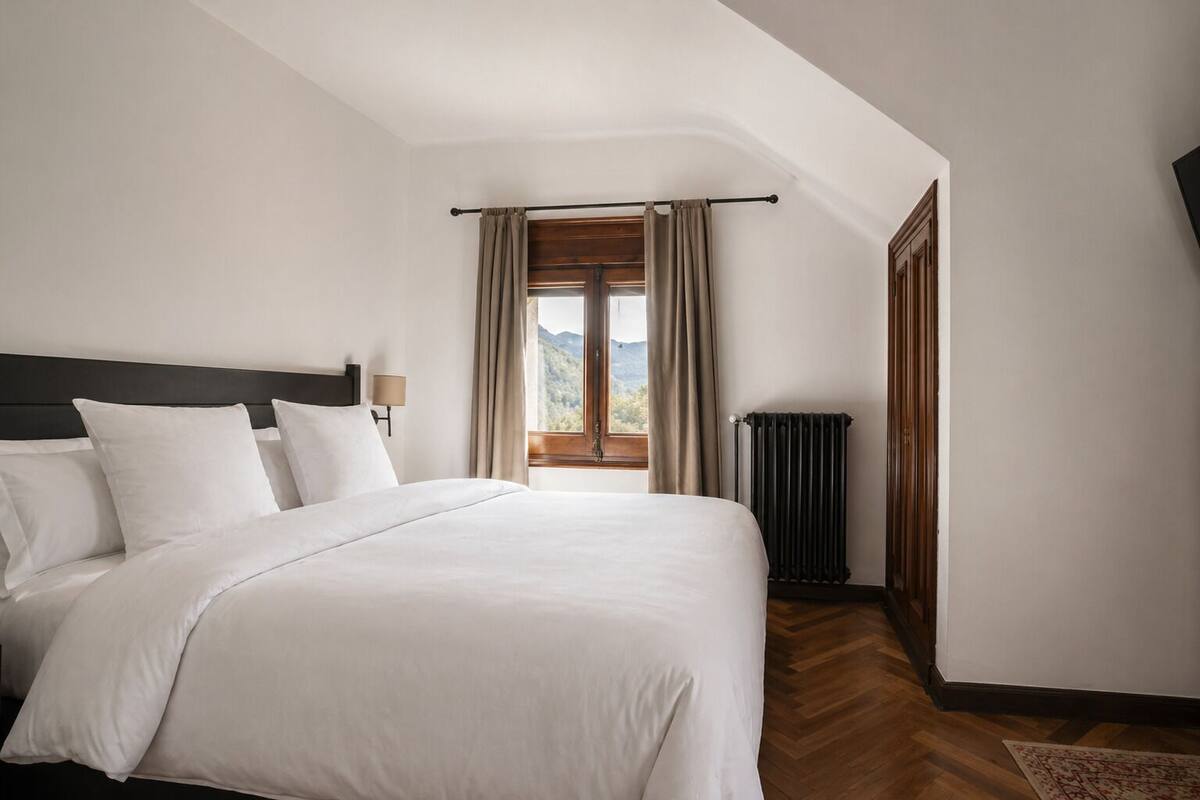 Een net opgemaakt bed met witte lakens, een houten hoofdbord, een nachtkastje met een lamp, een radiator en uitzicht op een bergachtig landschap door een raam met gordijnen.