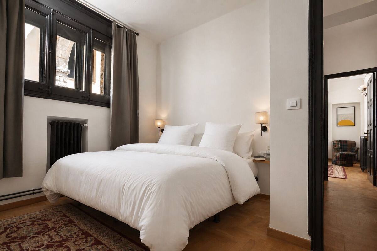 Een slaapkamer met een groot bed, twee bedlampen, een radiator en uitzicht op de straat door een raam.