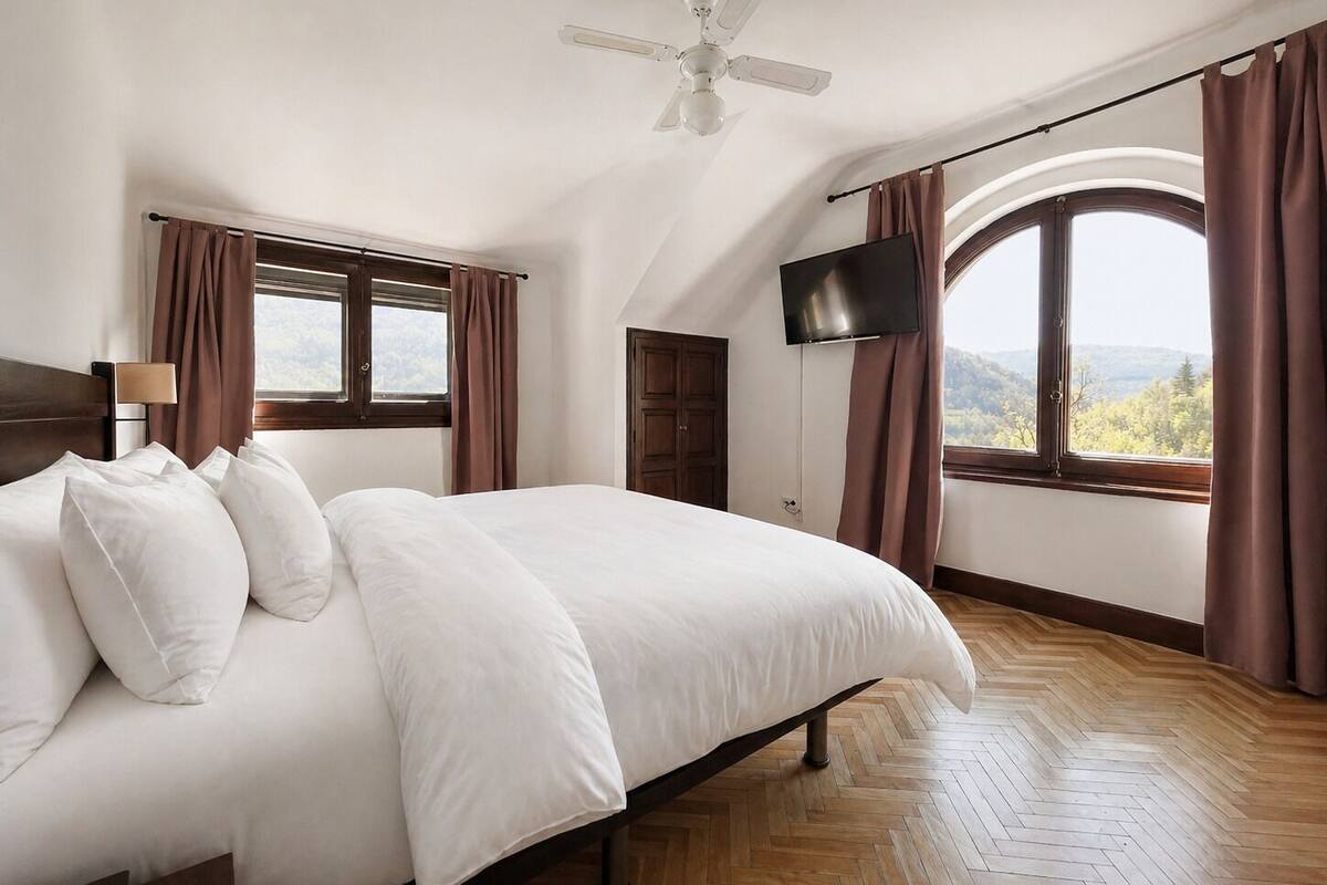 Een slaapkamer met een bed, een plafondventilator, een televisie en een raam met gordijnen.