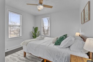 A bedroom with a bed, a ceiling fan, two windows, a plant, and a nightstand with a lamp.