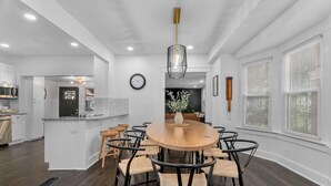 Een moderne keuken met witte kasten, een houten eettafel omringd door stoelen, en een groot raam met zonwering.