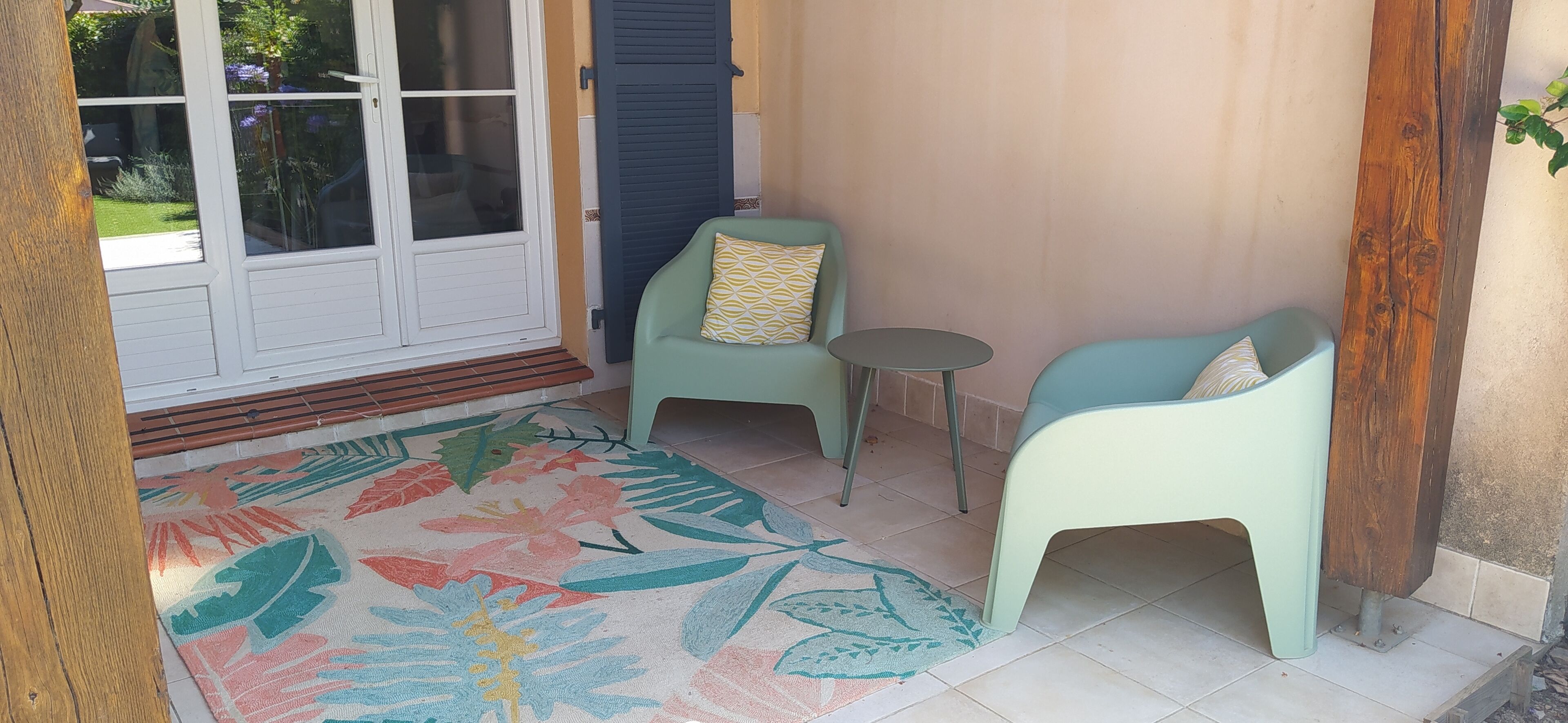 A sunlit room with a large window, a colorful floral rug, two light green chairs, a small round table, and a wooden bench.