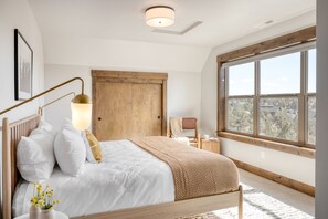 Ein Schlafzimmer mit großem Fenster, einer Holztür, einem Nachttisch mit Lampe und einem Bett mit weißer Bettwäsche und brauner Tagesdecke.