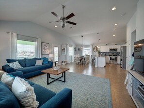 A spacious living room with a blue sofa, a wooden coffee table, and a ceiling fan. A kitchen with white cabinets and a dining area with a table and chairs are visible in the background.