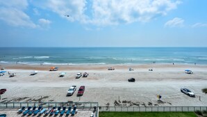 Une plage bordée d’une rangée de transats, un parking avec plusieurs voitures, et une vue dégagée sur l’océan.