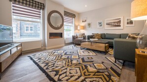 A modern living room with a sofa, coffee table, and a TV. There are large windows with blinds, a fireplace, and a patterned rug. - Howe Keld 5 First Floor (Keswick)