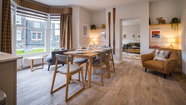 A modern dining area with a wooden table set for six, surrounded by chairs. A cozy armchair is visible in the background, and a window offers a view of the outside. - Howe Keld 5 First Floor (Keswick)