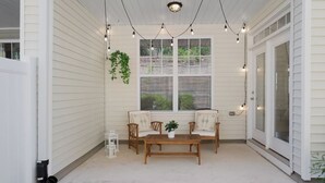 A covered porch with a wooden table and two chairs, string lights, and a hanging plant.