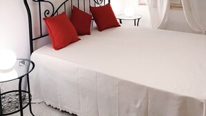 Un lit bien fait, recouvert d’une couvre-lit blanche et agrémenté de trois oreillers rouges, avec une tête de lit en métal noir et deux petites tables de chevet surmontées de lampes blanches.