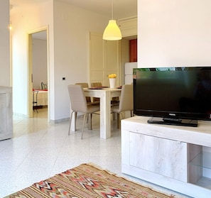 A modern living room with a flat-screen TV on a white cabinet, a dining area with a table and chairs, and a patterned rug on the floor.