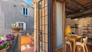 Una puerta abierta que da a un comedor con una mesa, sillas, una estantería y vistas al balcón exterior con plantas en macetas.