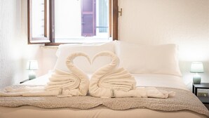 Habitación de hotel con cama, dos lámparas de noche, ventana con contraventanas y una disposición de toallas con forma de corazón sobre la cama.