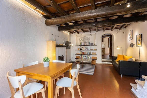Uma sala de estar aconchegante com uma mesa de jantar de madeira, cadeiras brancas, um sofá preto e uma estante de livros.