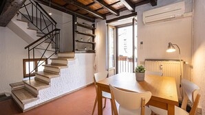 Una escalera con escalones de madera y barandillas negras, una mesa de comedor con sillas, una planta en maceta y un equipo de aire acondicionado en la pared.
