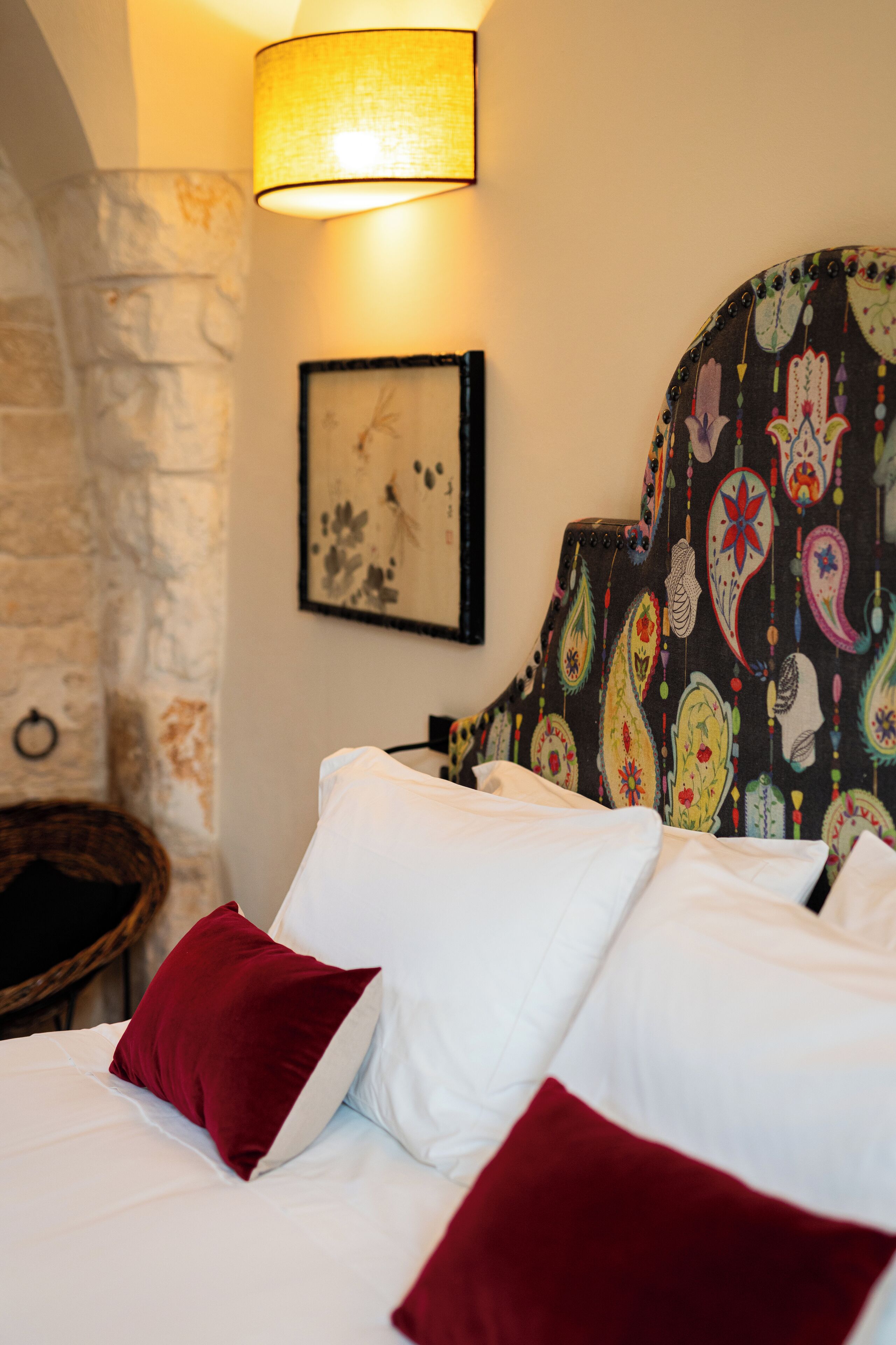 A bed with white and red pillows, a patterned headboard, a wall-mounted lamp, and a framed artwork.