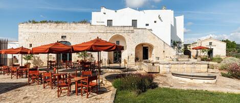 An outdoor seating area with stone tables and chairs, a fountain, and a white building with arches.