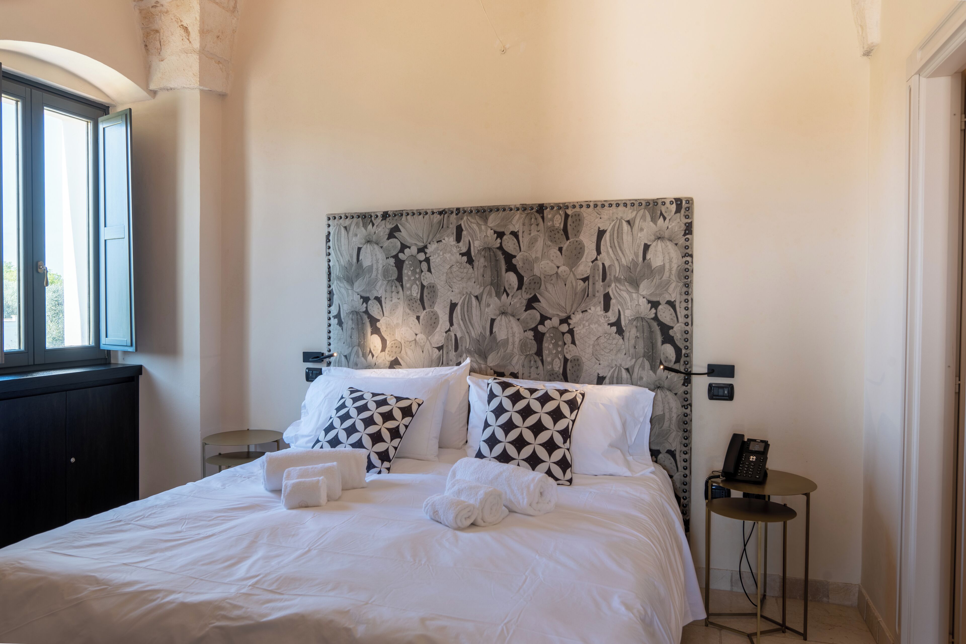 Un lit bien fait, avec du linge de lit blanc et deux coussins à motifs, une table de chevet avec un téléphone, et une fenêtre donnant sur un paysage verdoyant.