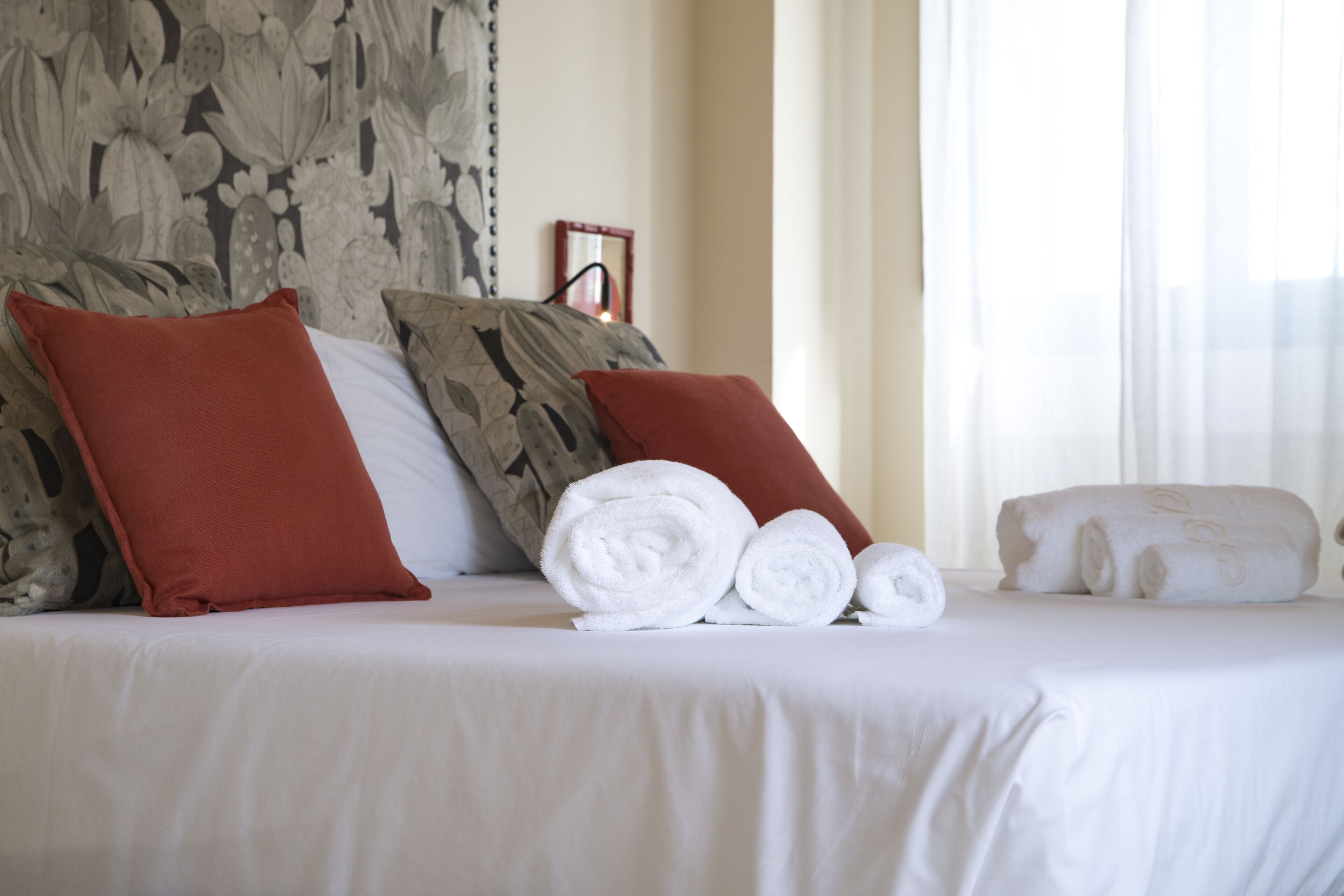 A neatly made bed with white linens, red and gray pillows, and rolled white towels.