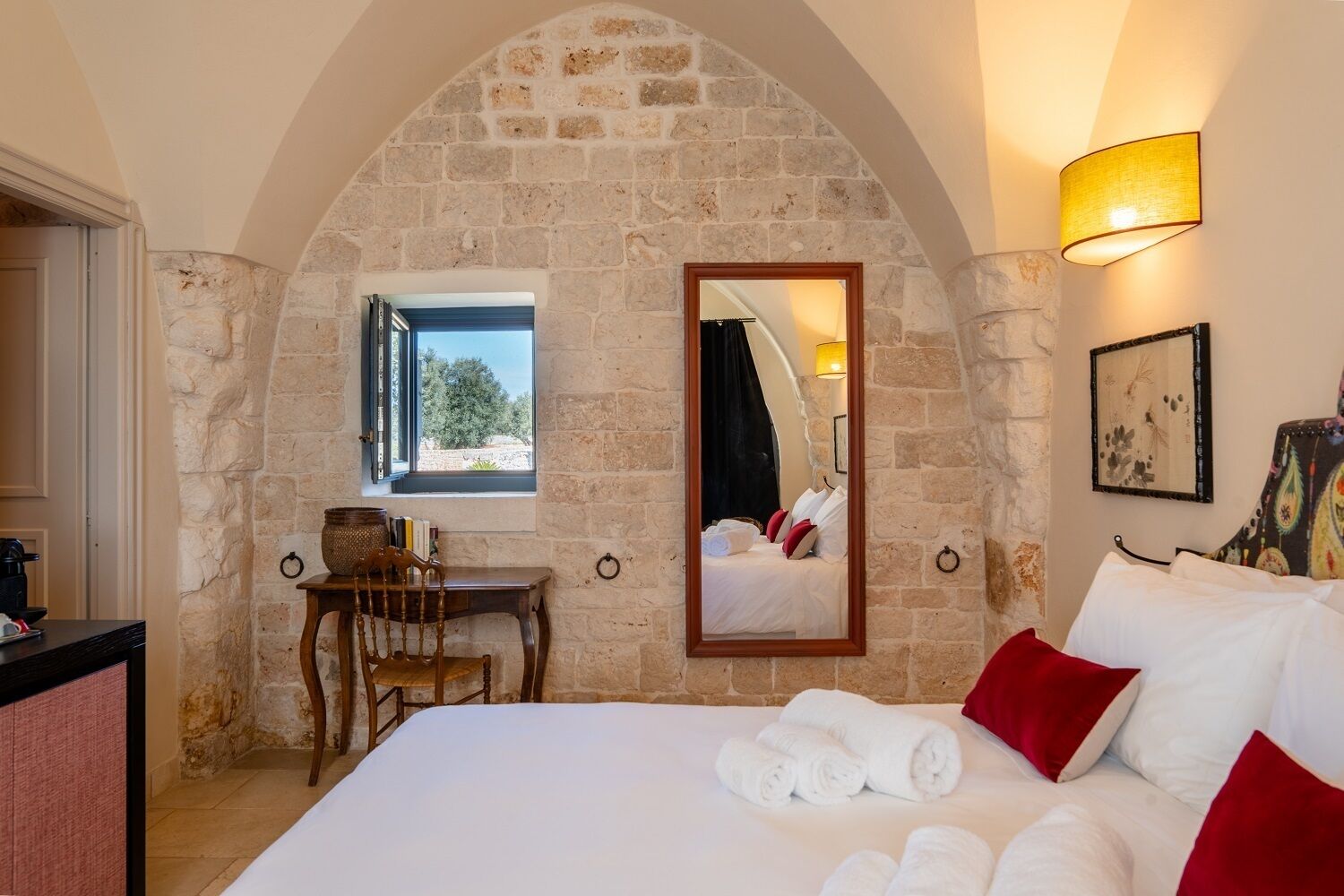 Un dormitorio con pared de piedra, cama con ropa blanca y almohadas rojas, escritorio con silla, ventana y espejo que refleja la cama.