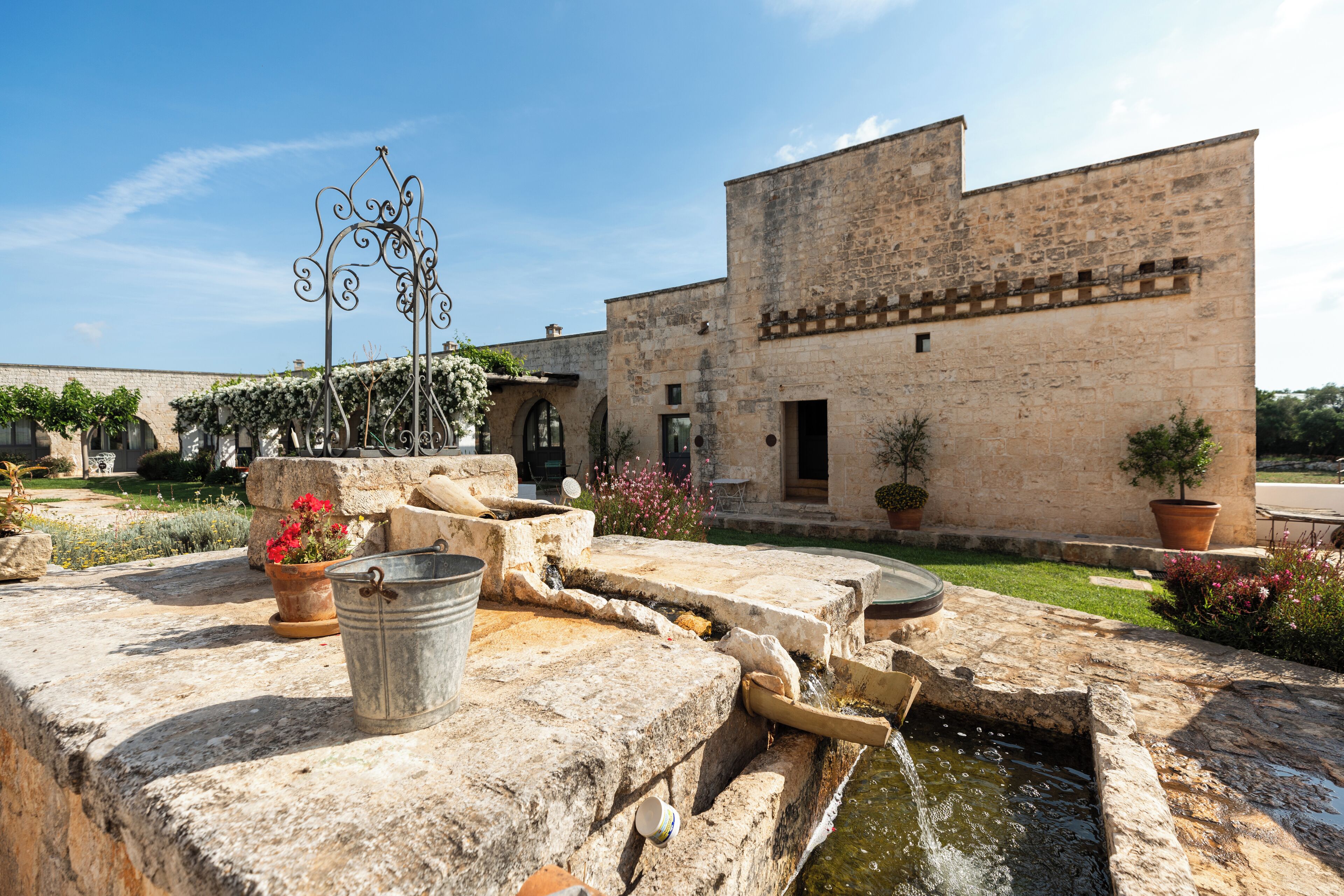 Un patio con un pozo de piedra, plantas en macetas y un edificio con techo de tejas.