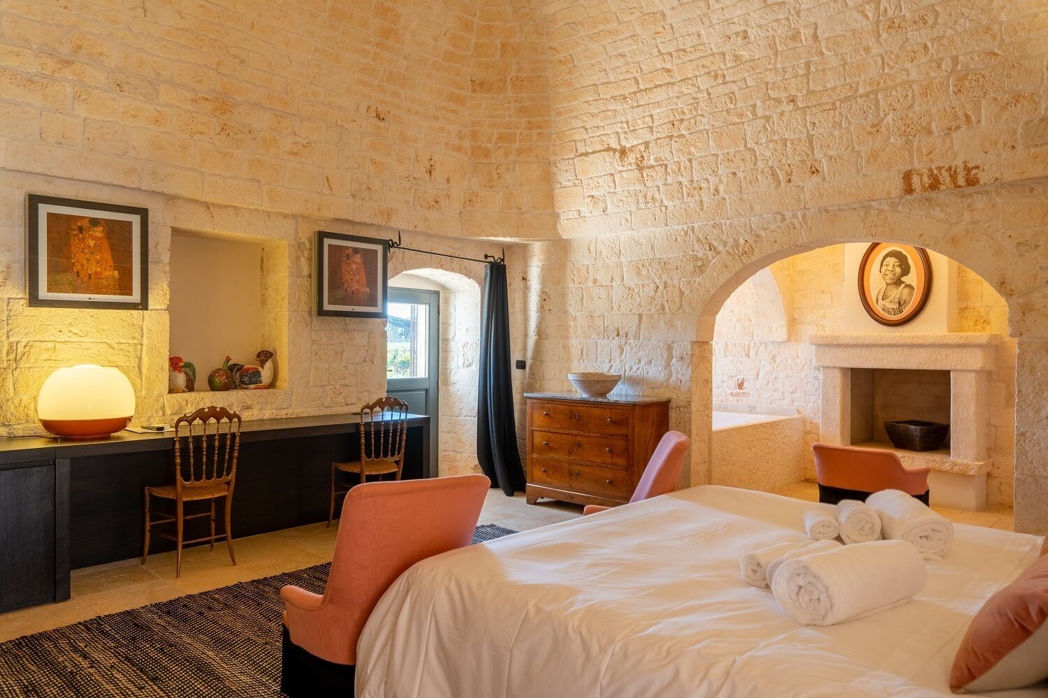A bedroom with a brick wall, a fireplace, a bed with rolled towels, a desk with a lamp, and a chair.
