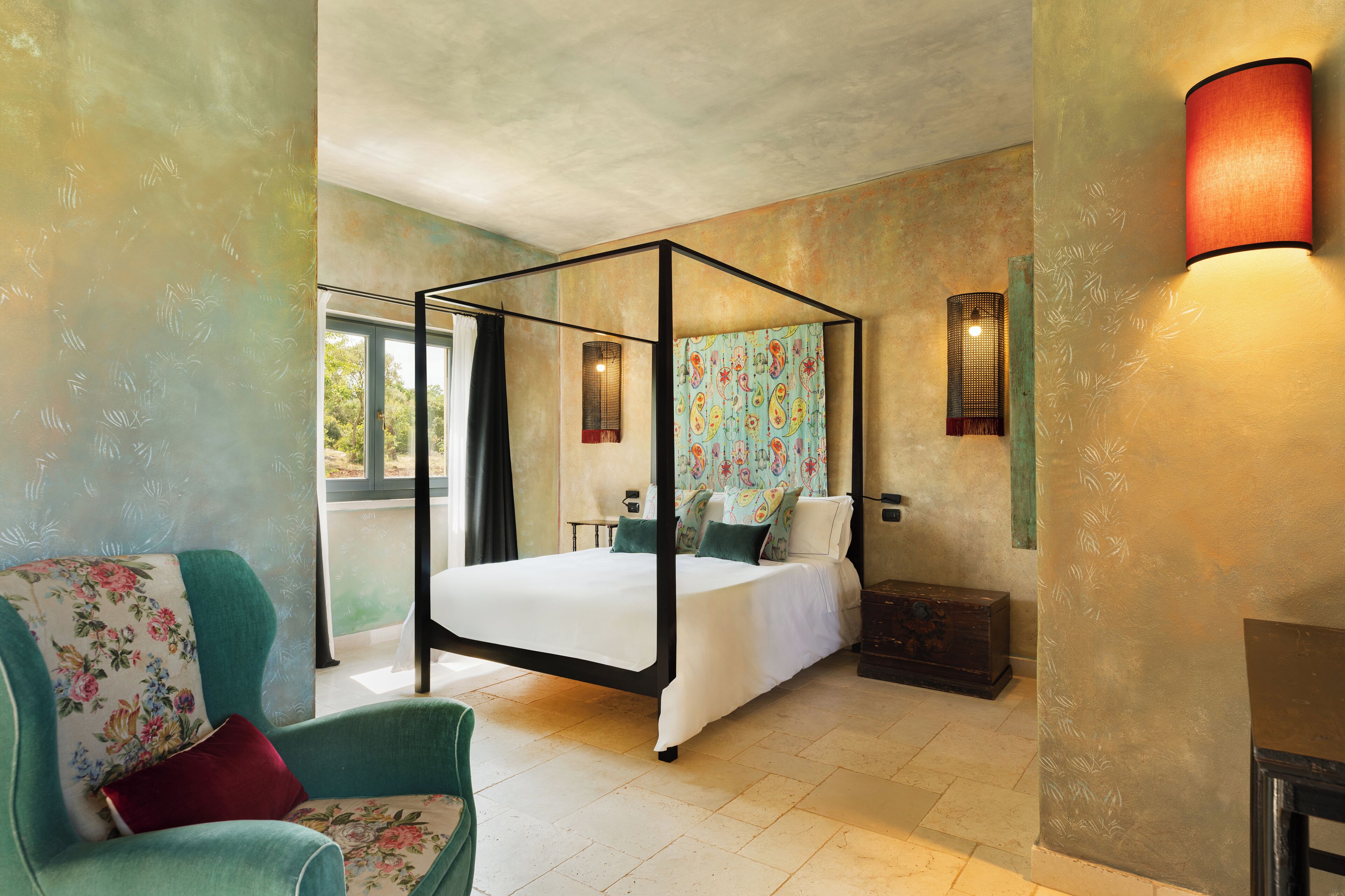 A four-poster bed with a canopy, a floral-patterned wall hanging, and a nightstand with a lamp.