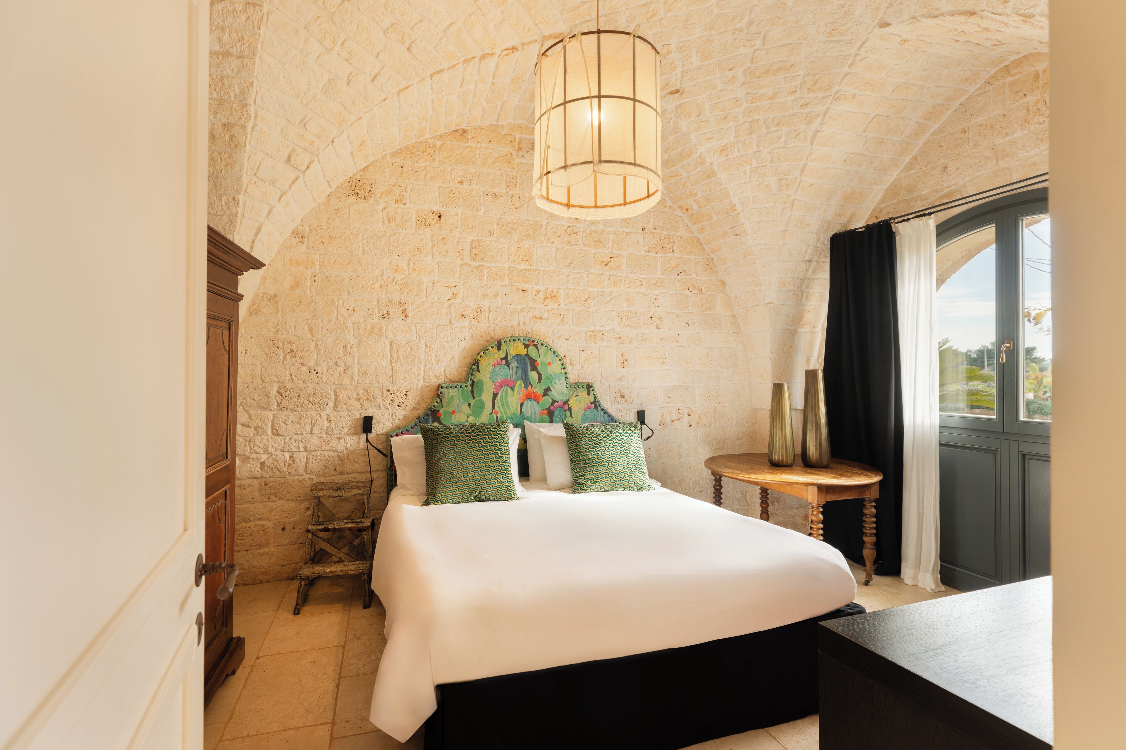 A bedroom with a stone wall, a bed with green pillows, a wooden chair, a small table, and a hanging light fixture.