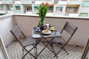 A balcony with a table set for two, a vase with flowers, and a view of a residential building.