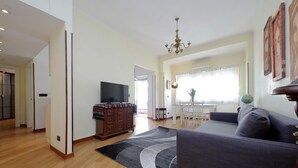 A spacious living room with a large grey sofa, wooden flooring, a flat-screen TV, a chandelier, and a dining area with a table and chairs.