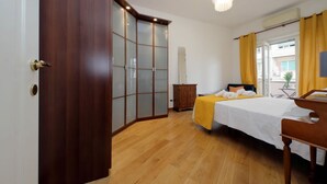 A hotel room with a wooden wardrobe, a bed with a yellow blanket, a wooden desk, and a window with curtains.
