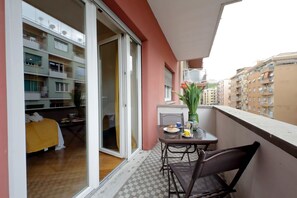 Ein Balkon mit Tisch und Stühlen, eine Glasschiebetür, die zu einem Raum mit einem Bett und einer gelben Decke führt.