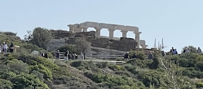 An ancient Greek temple with columns and steps, surrounded by greenery and people.