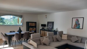 Moderni olohuone, jossa on kulmasohva, ruokailutila tuoleineen, televisio ja taideteoksia seinällä.