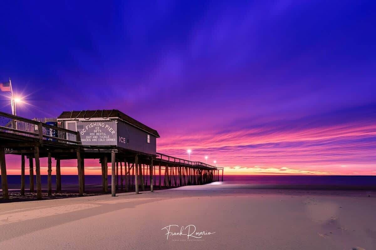 Eine Mole, die sich in einen Strand erstreckt, bei Sonnenuntergang, mit einem Gebäude, auf dem „Ice FISHER“ und „Ice Rental“ stehen.