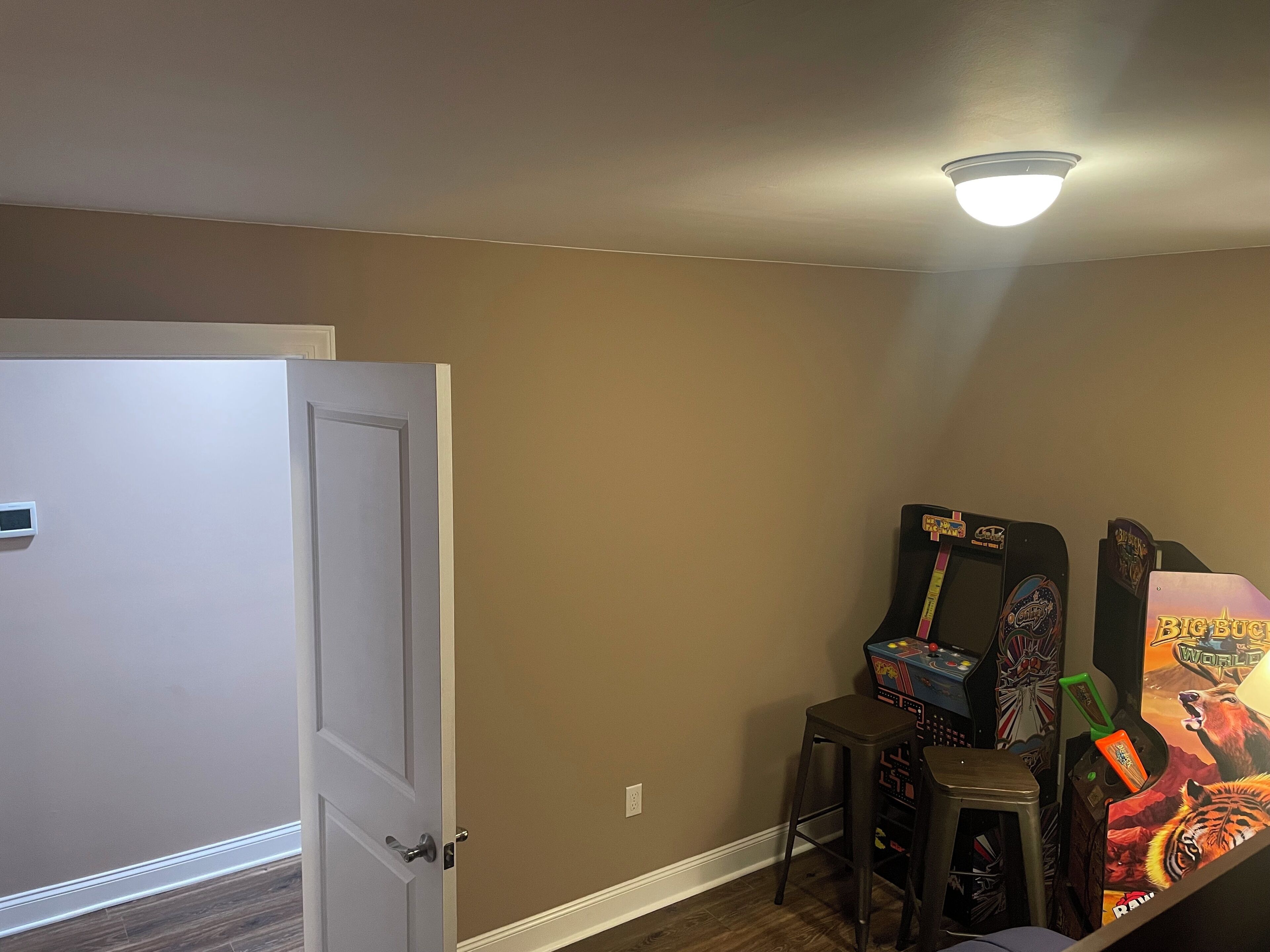 A room with beige walls, a white door, and a ceiling light. There are two stools and a colorful arcade machine.