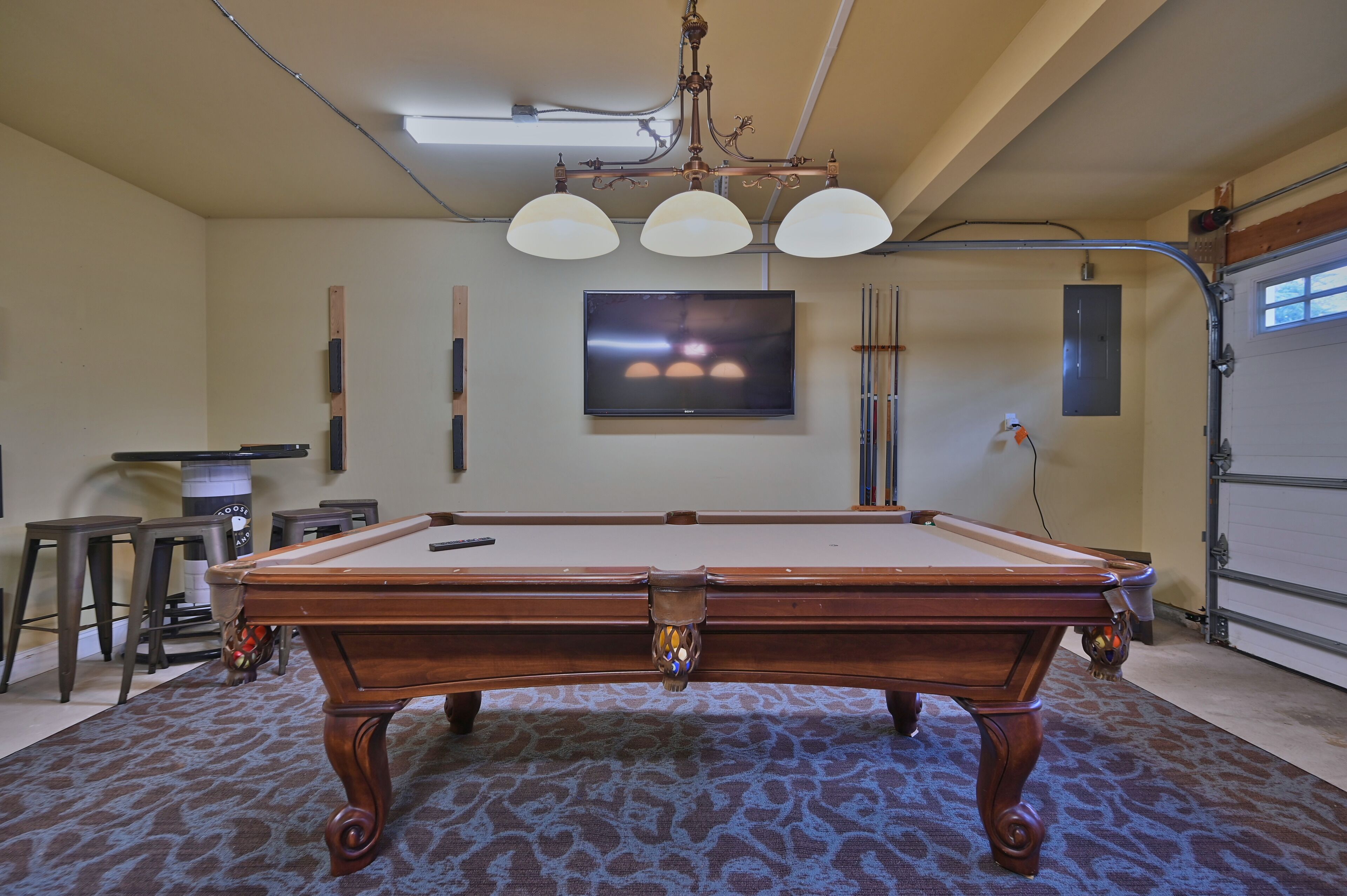 A pool table in a room with a patterned carpet, a mounted TV, and a chandelier.
