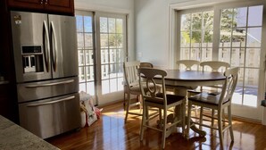 A kitchen with a stainless steel refrigerator, wooden cabinets, a dining table with white chairs, and large windows.